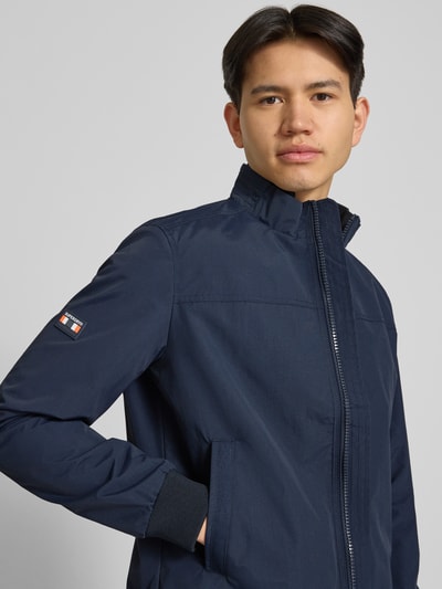 Männermodel trägt Jacke mit Stehkragen und Eingrifftaschen von Superdry in Dunkelblau, Regular-Fit, Nahaufnahme Oberkörper, Reißverschluss, Rückenlänge bei Größe M 70 cm, Schulterbreite 44 cm, Brustweite 41 cm, Ärmellänge 68 cm.