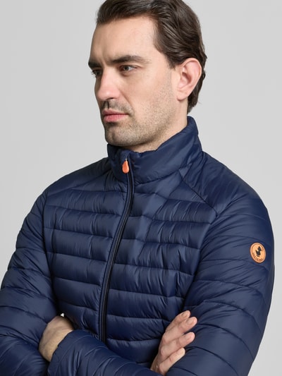 Männermodel trägt Steppjacke mit Zweiwegereißverschluss Modell 'Alexander' von SAVE THE DUCK in Blau, Nahaufnahme Oberkörper. Stepp-Optik, gefütterter Stehkragen, sichtbares Markenlogo, Regular Fit.