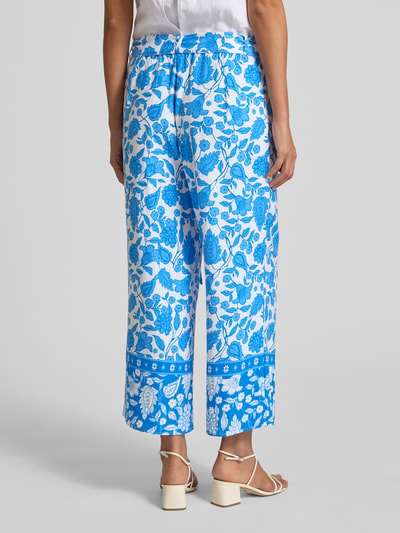 Frauenmodel trägt Straight Fit Stoffhose aus reiner Viskose von More & More mit floraler Allover-Print, elastischem Bund, Zierschleife, verkürzter Passform, blaue und weiße Farben, Ganzkörperansicht Rückansicht.