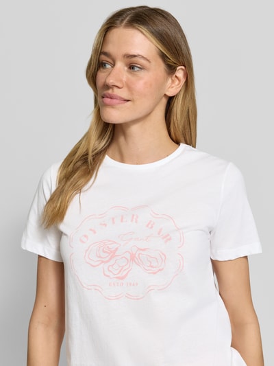 Frauenmodel trägt T-Shirt mit Motiv-Print und Rundhalsausschnitt von Gant, weiß, gerader Schnitt, Motiv in zartem Rosa, Nahaufnahme Oberkörper. Baumwoll-Mix mit Stretch-Anteil, Rückenlänge bei Größe S 58 cm.