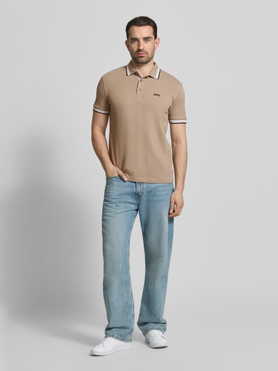 Męski model nosi Regular Fit Poloshirt aus reiner Baumwolle Modell 'PADDY' von BOSS Green w jednolitym kolorze beżowym, krój regular fit, bawełna pikowana, kołnierzyk polo, krótki rękaw, nadrukowane logo, zapięcie na trzy guziki, tekstura gładka, całopostaciowe ujęcie z przodu.