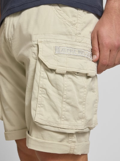 Mężczyzna nosi Shorts mit Cargotaschen Modell 'CREW SHORT' von Alpha Industries, bawełna z domieszką elastanu, krój regular fit, kolor beżowy, widoczna obszerna kieszeń cargo i naszywka z logo, szerokość w rozmiarze 32: 44 cm, długość nogawki wewnętrznej: 22 cm, detaliczne ujęcie tylnej części nogawki.