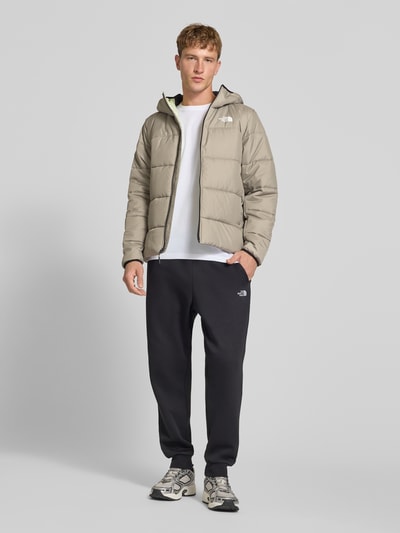 Männermodel trägt Regular Fit Jacke in Stepp-Optik von The North Face in Beige mit Kapuze, Logo-Print, Reißverschluss, Ganzkörperansicht von vorne. Kontext: Rückenlänge bei Größe M 68 cm, Schulterbreite 46 cm, Brustweite 55 cm, Ärmellänge 70 cm.