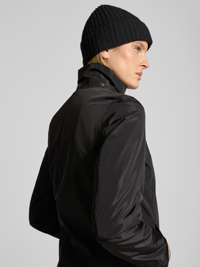 Frauenmodel trägt Regular Fit Mantel mit Logo-Applikation von NAUTGÅRD in Schwarz, glatte Textur, Stehkragen mit Logo-Detail, Eingrifftaschen, Kapuze mit Tunnelzug, Rückenlänge 88 cm, Schulterbreite 37 cm, Brustweite 48 cm, Ärmellänge 63 cm, Ganzkörperansicht Rückansicht.