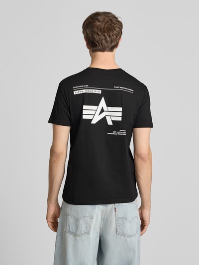 Männermodel trägt T-Shirt mit Label-Patch von Alpha Industries in Schwarz, Reine Baumwolle, Gerader-Schnitt, Rückenlänge 66 cm, Ganzkörperansicht Rückansicht.