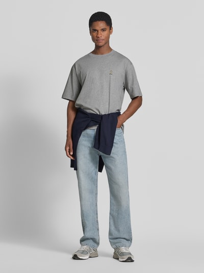Męski model nosi szarą koszulkę krój oversized Relaxe Fit Oversized Shirt aus reiner Baumwolle model 'ICONIC Å MAARKOS' od ARMEDANGELS, jasnoniebieskie jeansy, Ganzkörperansicht von vorne. Koszulka Herren Oversized Shirt od ARMEDANGELS.