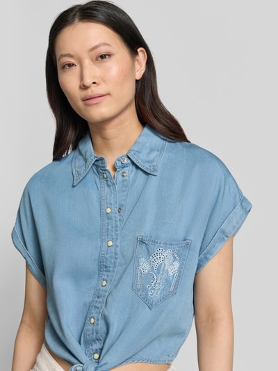 Modelka nosi bluzkę jeansową z lyocellu, krój loose fit, model 'AMBRA' od Guess, kołnierzyk typu kent, listwa guzikowa, krótkie rękawy z zakładką, brustasza z haftem, wiązany detal, długość tyłu w rozmiarze S 47 cm, długość rękawów w rozmiarze S 8 cm, jasnoniebieski kolor, miękka tekstura, zbliżenie na górną część ciała, widok z przodu.