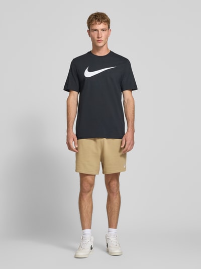 Männermodel trägt Regular Fit Sweatshorts aus Baumwoll-Mix von Nike in Beige, kombiniert mit dunklem Shirt und weißen Sneakern, Ganzkörperansicht von vorne. Herren Sweatshorts von Nike.