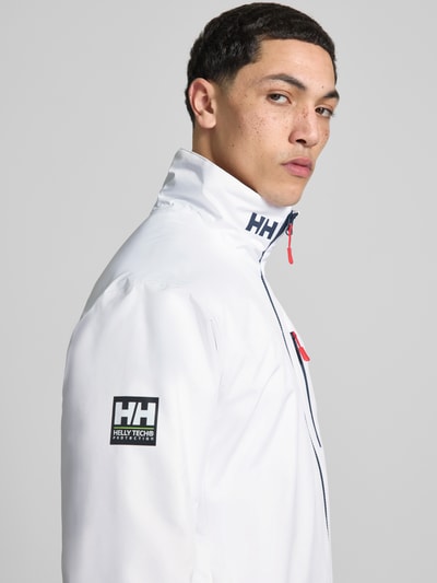 Männermodel trägt Midlayer Jacke mit Stehkragen Modell 'CREW' von Helly Hansen in Weiß mit Logo-Print, Label-Patch, Reißverschluss; Nahaufnahme Oberkörper. Gerader-Schnitt, Brusttasche mit Reißverschluss, seitliche Reißverschlusstaschen, Material glatte Textur. Rückenlänge 74 cm, Schulterbreite 49 cm, Brustweite 52 cm, Ärmellänge 68 cm bei Größe M.