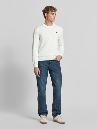 Männermodel trägt weißen Regular Fit Pullover aus Baumwoll-Mix von Lacoste, kombiniert mit blauen Jeans, Ganzkörperansicht von vorne. Herren Pullover von Lacoste.