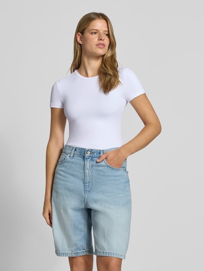 Vrouwenmodel draagt Slim Fit T-Shirt met ronde hals model 'LULA' van PIECES, dames t-shirt, wit met gladde textuur, blauwe denim short, driekwartvooraanzicht.