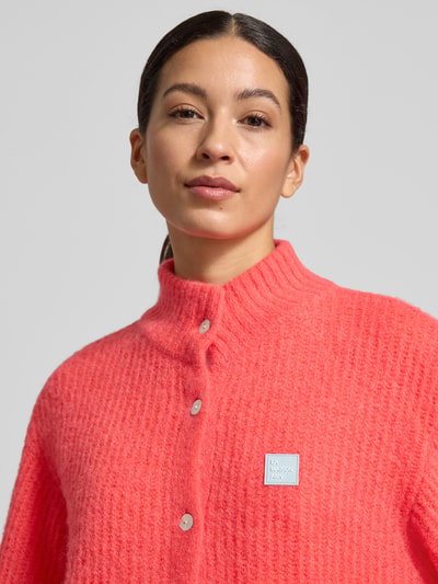 Frauenmodel trägt Strickjacke mit Knopfleiste von American Vintage in Koralle, gerippte Abschlüsse, Stehkragen, Material-Mix mit Alpaka, Nahaufnahme Oberkörper.