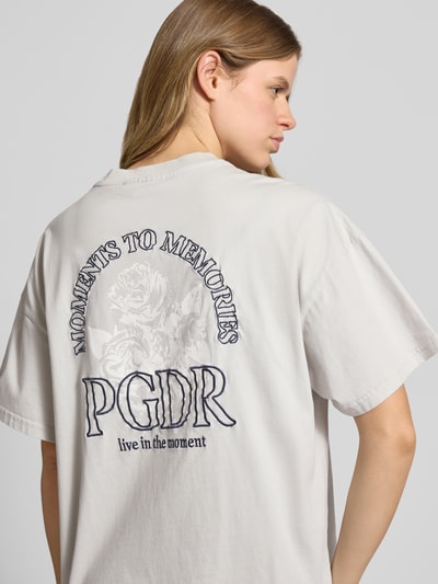 Modelka nosi Oversized T-Shirt mit Label-Stitching Modell 'MARIALVA' od Pegador, czysta bawełna, kolor jasnoszary z czarnym nadrukiem na plecach, okrągły dekolt, długość tyłu w rozmiarze S: 61 cm, ujęcie z tyłu, zbliżenie na górną część pleców.