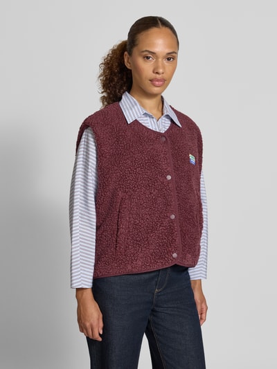 Frauenmodel trägt Regular Fit Weste aus Teddyfell Modell 'Hoktown' von American Vintage in Dunkelrot mit Logo-Patch, Ganzkörperansicht von vorne. Damen Weste mit Knopfleiste, Rückenlänge 57 cm, Schulterbreite 43 cm, Brustweite 63 cm, Teddyfell-Textur, lockerer Schnitt, gestreifte Bluse und dunkle Jeans.