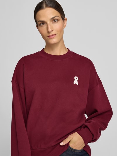 Frauenmodel trägt Oversized Sweatshirt aus reiner Baumwolle Modell 'ALIZAA' von ARMEDANGELS in Dunkelrot mit weißem Logo, Nahaufnahme Oberkörper.