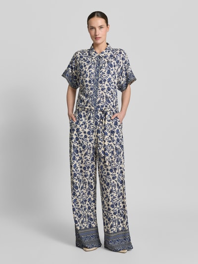 Vrouwenmodel draagt jumpsuit uit zuiver viscose met all-over look, overhemdkraag en strikceintuur, model 'Mathilde' van LOLLYS LAUNDRY, relaxed fit, ruglengte maat S 46 cm, borstwijdte 44 cm, mouwlengte 17 cm, totale lengte 160 cm, binnenbeenlengte 77 cm, kleuren blauw en crème, geometrische en florale vormen, gladde textuur, model van voren volledig zichtbaar, ganzebodyshot.
