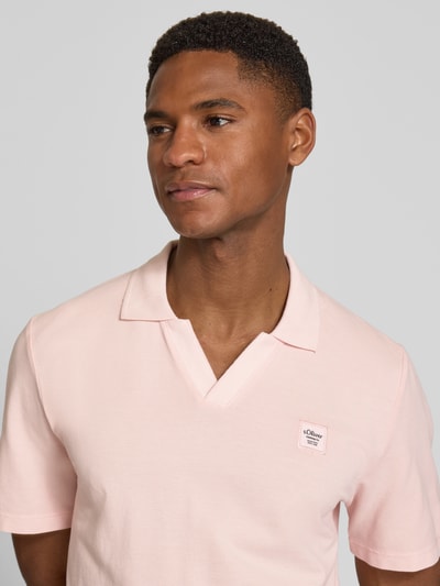 Mężczyznamodel nosi Regular Fit Poloshirt aus reiner Baumwolle von s.Oliver RED LABEL w jednolitym różowym kolorze, tekstura piké, aplikacja label patch, krój regular fit, kołnierz polo, dekolt w serek, krótkie rękawy z prążkowanym mankietem, rozcięcia po bokach na dole, materiał czysta bawełna, zbliżenie na górną część ciała.