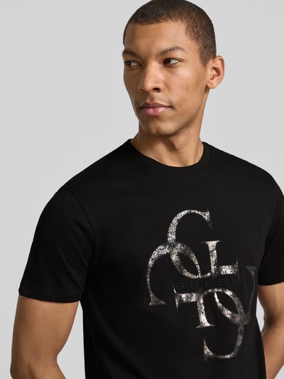 Mężczyzna nosi T-Shirt mit Label-Detail od Guess, czarna bawełna, prosty krój, nadruk z logo, długość tyłu w rozmiarze M: 69 cm, zbliżenie na górną część ciała