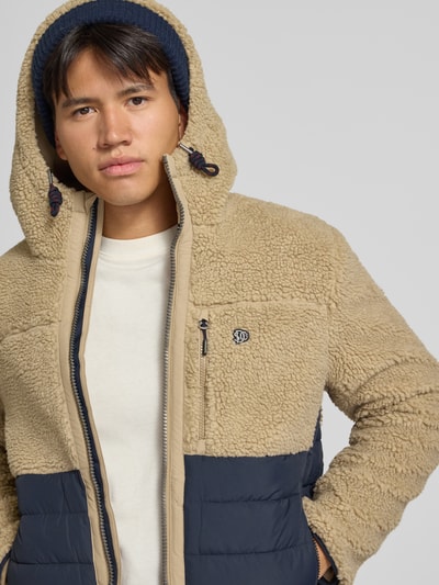 Männermodel trägt Regular Fit Jacke mit Teddyfell Modell 'Borg' von Superdry, zweifarbige Optik, Kapuze, Reißverschluss, Nahaufnahme Oberkörper. Oni widzi kurtkę damską z teddy, krój regular fit, model 'Borg', kieszenie napoleońskie i wsuwane, kaptur ze ściągaczem, długość tyłu w rozmiarze M: 64 cm, szerokość ramion w rozmiarze M: 45 cm, szerokość klatki piersiowej w rozmiarze M: 55 cm, długość rękawa w rozmiarze M: 68 cm.