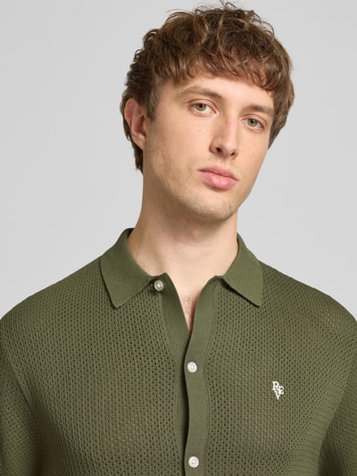Männermodel trägt Slim Fit Strick-Poloshirt mit Label-Stitching von REVIEW, olivgrün, Viskose-Mix, Kontraststreifen, Nahaufnahme Oberkörper.