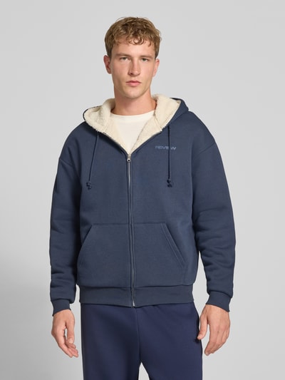 Männermodel trägt Regular Fit Sweatjacke aus Baumwoll-Mix mit Teddyfutter von REVIEW, dunkelblau, Logo-Detail, Ganzkörperansicht von vorne.