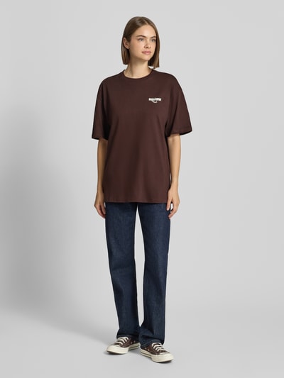 Frauenmodel trägt Oversized Fit T-Shirt aus reiner Baumwolle mit Label-Detail von REVIEW in Dunkelbraun, kombiniert mit Jeans und Sneakern, Damen T-Shirt mit Print auf der Rückseite, Rückenlänge bei Größe S: 69 cm, Ganzkörperansicht von vorne.