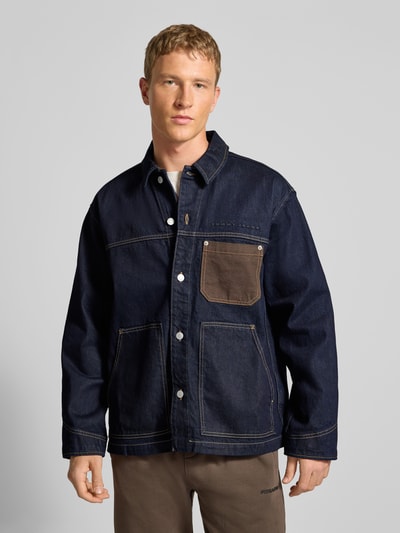 Männermodel trägt Regular Fit Jeansjacke aus reiner Baumwolle von Tommy Jeans in Melange-Optik, dunkles Denim, Kontrast-Brusttasche, Nahaufnahme Oberkörper.