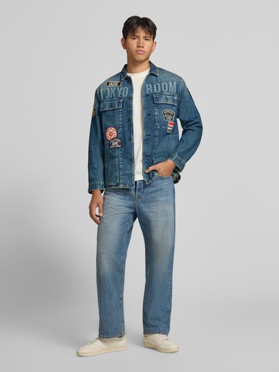 Männermodel trägt Regular Fit Jeansjacke aus Baumwoll-Mix von Replay in Blau mit Allover-Patches und Knopfleiste, Ganzkörperansicht von vorne.