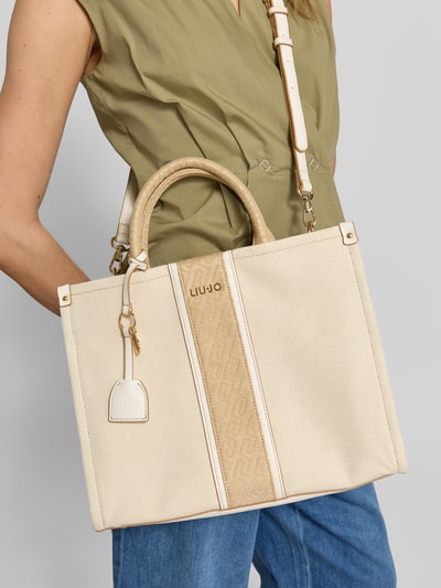 Frauenmodel trägt Handtasche mit Tragehenkeln und Schulterriemen von Liu Jo White aus Reine Baumwolle, Label-Applikation, Hauptfach mit Reißverschluss, Breite 34 cm, Länge 27 cm, Tiefe 12 cm, Riemenlänge 31 cm, Creme- und Beigetöne, strukturierte Oberfläche, Ganzkörperansicht von vorne.