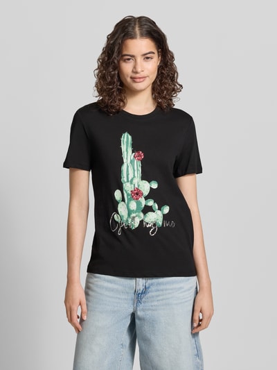 Frauenmodel trägt Damen T-Shirt von Only, Slim Fit Modell 'KITA', Optik: Unifarben, Motiv-Print mit Pailletten, Rundhals, Kurzarm, Jersey aus reiner Baumwolle, Motiv zeigt grünen Kaktus mit rosa Blüten auf schwarzem Grund, ganzkörperansicht von vorne.