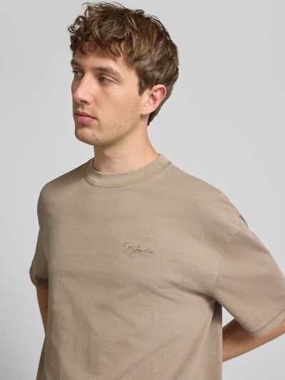 Męski model nosi Oversized T-Shirt mit Label-Stitching od Pegador w kolorze beżowym, czysta bawełna, krój oversize, okrągły dekolt, widoczne logo, długość tyłu w rozmiarze M: 70 cm, nahaufnahme górnej części ciała.