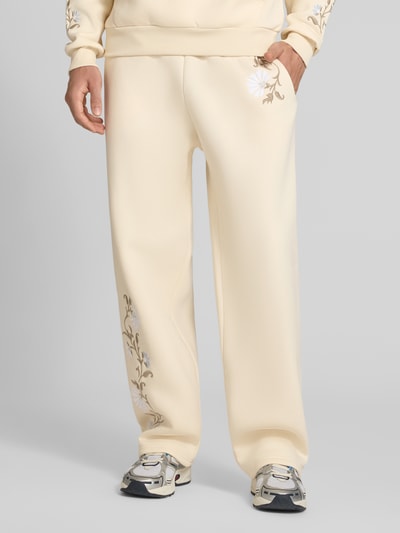Männermodel trägt Straight Fit Sweatpants mit floraler Stickerei von REVIEW in Beige mit elastischem Bund, Ganzkörperansicht von vorne.