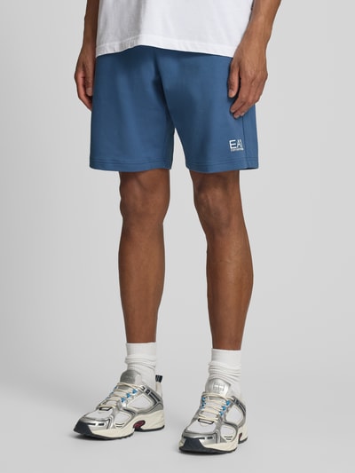 Männermodel trägt Sweatshorts mit Logo und Eingrifftaschen von EA7 Emporio Armani in Blau, Reine Baumwolle, Regular-Fit, Nahaufnahme Beine, Ganzkörperansicht von vorne.