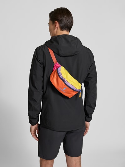 Männermodel trägt Bauchtasche mit Logo-Print Modell 'Bataan' von Cotopaxi in Orange, Gelb und Pink mit verstellbarem Schulterriemen, Hauptfach mit Reißverschluss, Ganzkörperansicht Rückansicht. Breite 23 cm, Länge 14 cm, Tiefe 11 cm, Riemenlänge 50 cm.