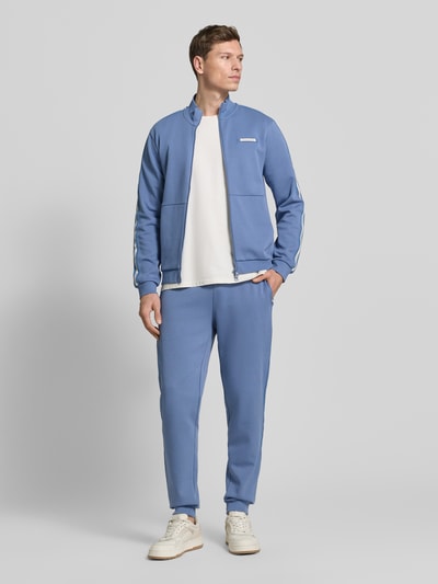 Männermodel trägt Sweatjacke aus Baumwoll-Mix von EA7 Emporio Armani in Blau mit Galonstreifen, Label-Detail, Zweiwege-Reißverschluss, Gerade Passform, elastische Abschlüsse, Känguru-Tasche, Ganzkörperansicht von vorne.