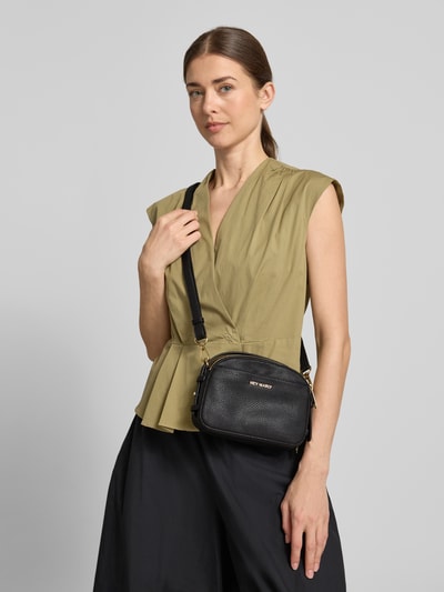 Frauenmodel trägt Umhängetasche mit Label-Detail Modell 'Style Mate' von Hey Marly in Schwarz, genarbte Textur, Hauptfach mit Reißverschluss, verstellbarer Schultergurt, Reißverschlussfach innen, Breite 21 cm, Länge 14 cm, Tiefe 8 cm, Länge Schulterriemen 44 cm, Ganzkörperansicht von vorne.