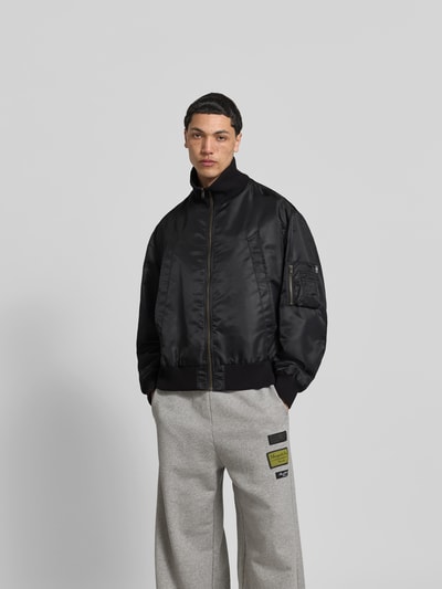 Männermodel trägt Regular Fit Bomberjacke mit Stehkragen von MM6 Maison Margiela, Schwarz, Eingrifftaschen, Ärmeltasche, Reißverschluss, elastische Abschlüsse, Rückenlänge 63 cm, Schulterbreite 54 cm, Ärmellänge 67 cm, Ganzkörperansicht von vorne.