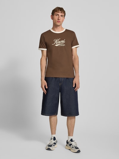 Męski model nosi T-Shirt mit Label-Print und Rundhalsausschnitt von Karl Kani z bawełny, krój regular fit, długość tyłu w rozmiarze M: 68 cm, widok całej sylwetki z przodu.