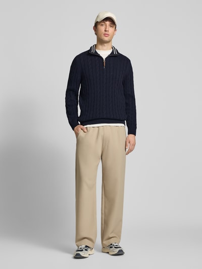 Männermodel trägt Strickpullover aus reiner Baumwolle von Superdry, Dunkelblau mit Zopfmuster, Troyer-Kragen, Label-Detail, eng anliegende Passform, Gerippte-Abschlüsse, Rückenlänge 70 cm, Ärmellänge 71 cm, Herren, Ganzkörperansicht von vorne.