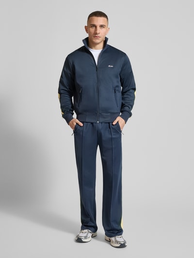 Männermodel trägt Track Pants mit Reißverschlusstaschen und Kordelzug von ANOTHER COTTON LAB, navyblau mit Gelbdetails, Wide Fit, Material-Mix mit Stretch, elastischer Bund, Ganzkörperansicht von vorne.
