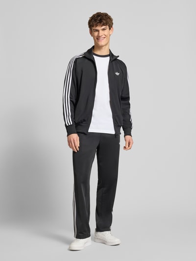 Männermodel trägt schwarze Sweatjacke mit Reißverschluss von adidas Originals mit weißen Streifen, Regular Fit, Ganzkörperansicht von vorne.