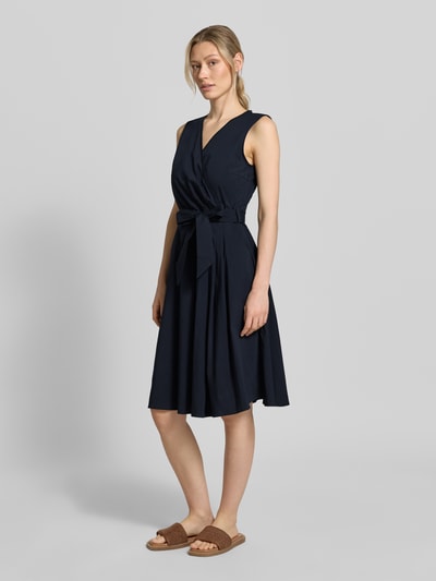 Frauenmodel trägt Kleid mit V-Ausschnitt in Wickel-Optik von More & More in Dunkelblau, Baumwoll-Mix, Tailliert, Ärmelloses-Design, Abnehmbares Taillenband, Ganzkörperansicht von vorne.