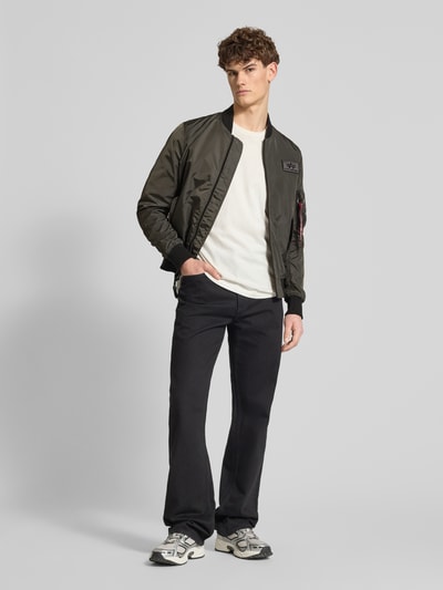 Mężczyzna model nosi Regular Fit Bomberjacke mit Logo-Patch od Alpha Industries w kolorze khaki, wpuszczane kieszenie, zamek błyskawiczny, kieszeń na rękawie; długość tyłu w rozmiarze M: 65 cm, szerokość ramion: 45 cm, szerokość w rozmiarze M: 54 cm, długość rękawów: 69 cm. Cała sylwetka z przodu.