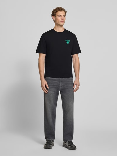 Männermodel trägt Regular Fit T-Shirt aus reiner Baumwolle Modell 'VIBING COCO' von REVIEW, schwarze Farbe, Ganzkörperansicht von vorne, Print auf der Rückseite, Rückenlänge bei Größe M 68 cm.