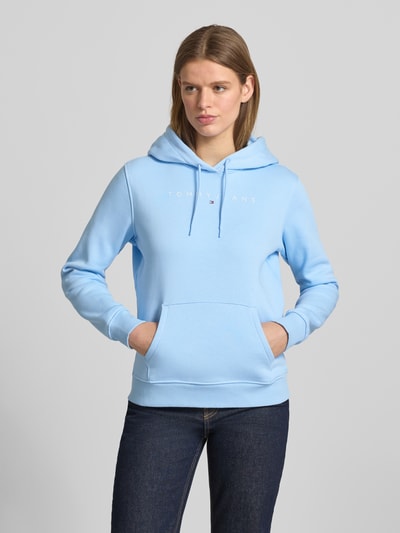Frauenmodel trägt Regular Fit Hoodie aus Baumwoll-Mix von Tommy Jeans in Hellblau, unifarben, langarm, Label Print, Sweat-Textur, Ganzkörperansicht von vorne.