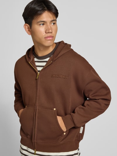 Männermodel trägt braune Sweatjacke mit Kapuze von ANOTHER COTTON LAB aus elastischem Baumwoll-Mix, lockerer Schnitt, Ganzkörperansicht von vorne.