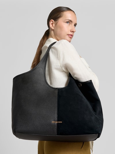 Frauenmodel trägt Tote Bag mit Logo-Applikation Modell 'Henly' von MICHAEL Michael Kors in Schwarz, Strukturmix, Ganzkörperansicht Rückansicht. Geräumige Form mit Hauptfach, Magnetverschluss, Schulterriemen, Innenfach mit Reißverschluss, Maße: 51x32x14 cm, Riemen 55 cm.