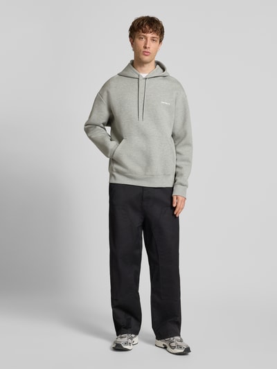 Męski model nosi Hoodie mit Kapuze od Carhartt Work In Progress, bawełniana mieszanka, luźny krój, szara, kangurka, logo, prążkowane wykończenia, widok całościowy z przodu.