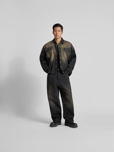 Männermodel trägt Relaxed Fit Jeanshemd im Used-Look von VETEMENTS aus reiner Baumwolle mit durchgehender Knopfleiste, Brusttaschen, langärmlig, Rückenlänge 74 cm, Schulterbreite 50 cm, Ärmellänge 74 cm, dunkle Farbverläufe, Ganzkörperansicht von vorne.