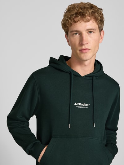 Männermodel trägt Regular Fit Hoodie aus Baumwoll-Mix Modell 'Soho' von JACK & JONES in Dunkelgrün mit Kapuze und Känguru-Tasche, Nahaufnahme Oberkörper. Der Hoodie zeigt elastische Abschlüsse, Logo-Aufdruck, Teil des Kontextes: Rückenlänge 66 cm, Ärmellänge 65 cm.